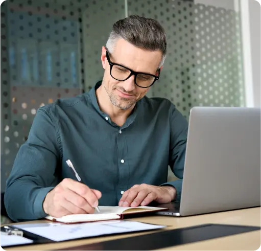 A man wearing glasses is focused on writing in a notebook, capturing his thoughts with a pen.