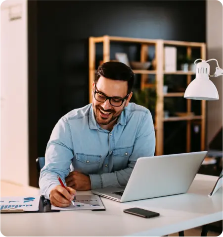 A smiling man wearing glasses is focused on his work while using a laptop.