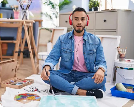 A man sits on the floor, wearing headphones and holding a paintbrush, immersed in his creative process.