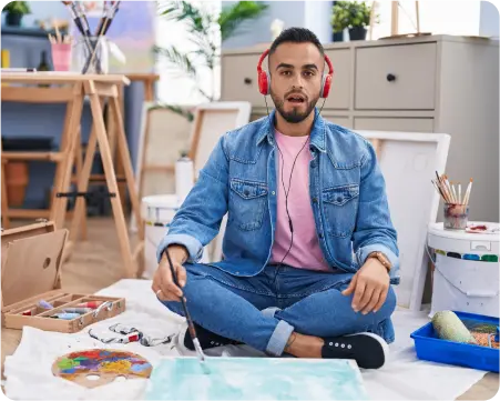 A man sits on the floor, wearing headphones and holding a paintbrush, immersed in his creative process.