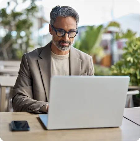 A man wearing glasses and a suit is seated at a table, working on a laptop.