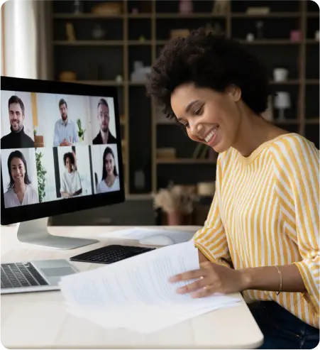 A woman smiles while seated at a desk, working on a laptop with a computer screen in front of her.