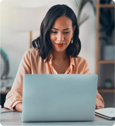 A woman seated at a desk, focused on her laptop, surrounded by a tidy workspace.