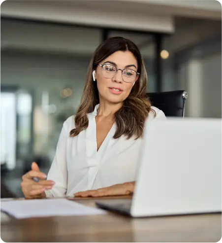 A woman wearing glasses is seated at a desk, focused on her laptop while working.