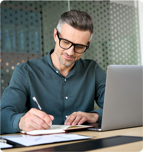 A man wearing glasses is writing in a notebook while simultaneously using a laptop at a desk.