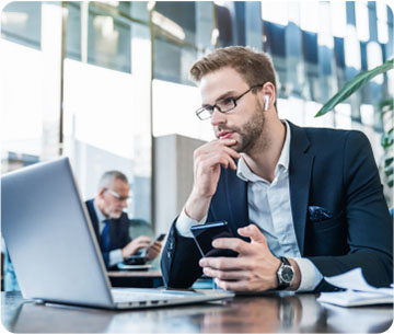 A man in a suit and glasses sits at a desk, focused on his phone while engaged in work.