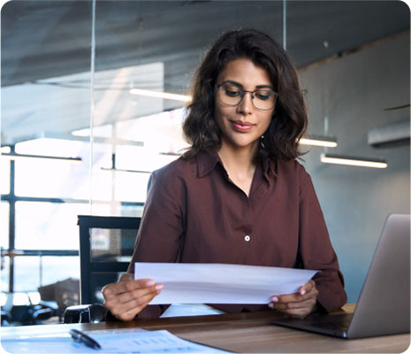 A woman wearing glasses sits at a desk, holding a piece of paper, focused on her work.