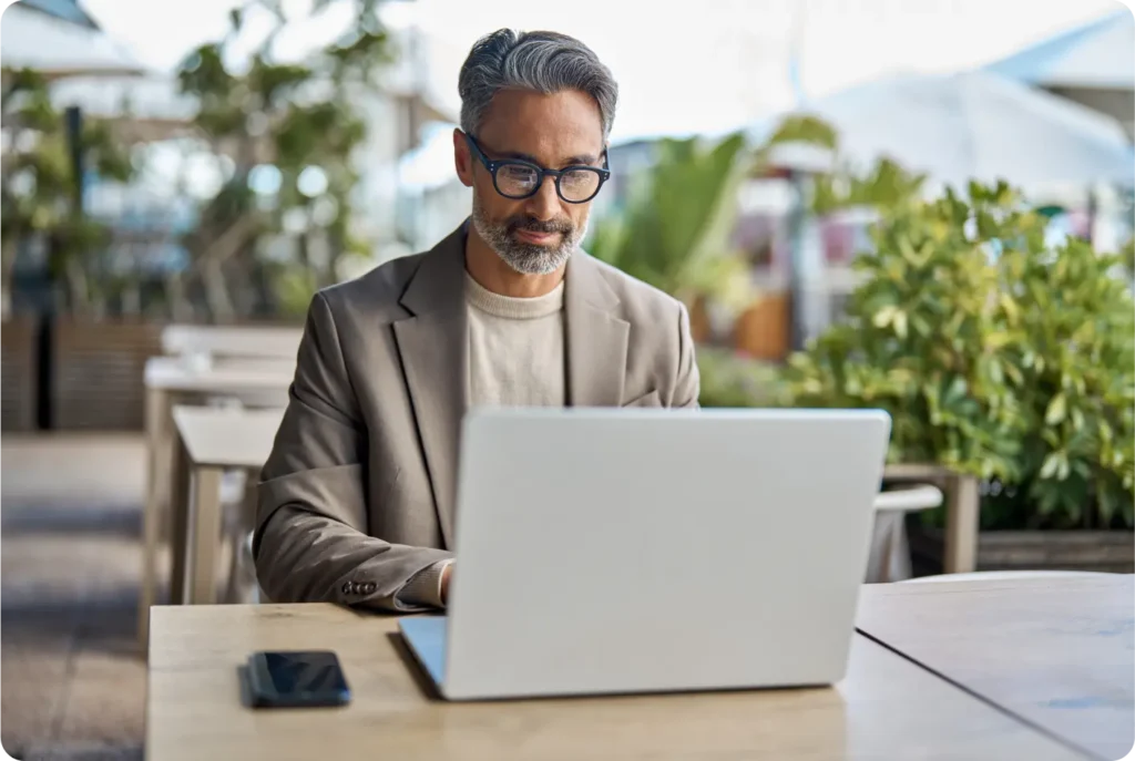 A man wearing glasses is seated at a table, focused on his laptop while working or studying.