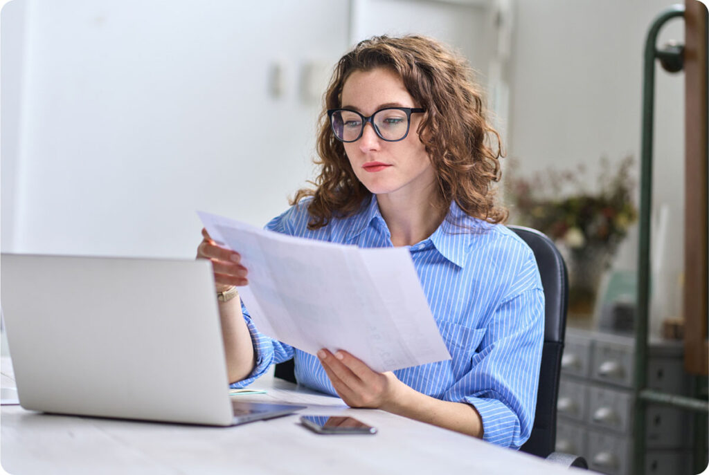 A woman wearing glasses intently examines a document displayed on her laptop screen.